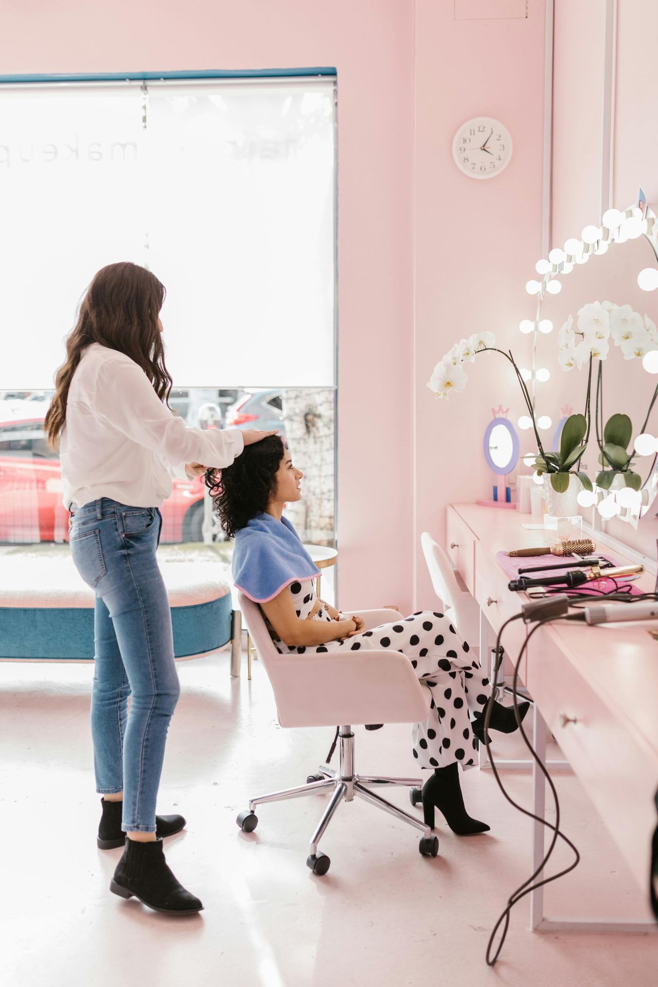 Hairdresser styling a woman's hair in a salon with pink decor and vanity lights.