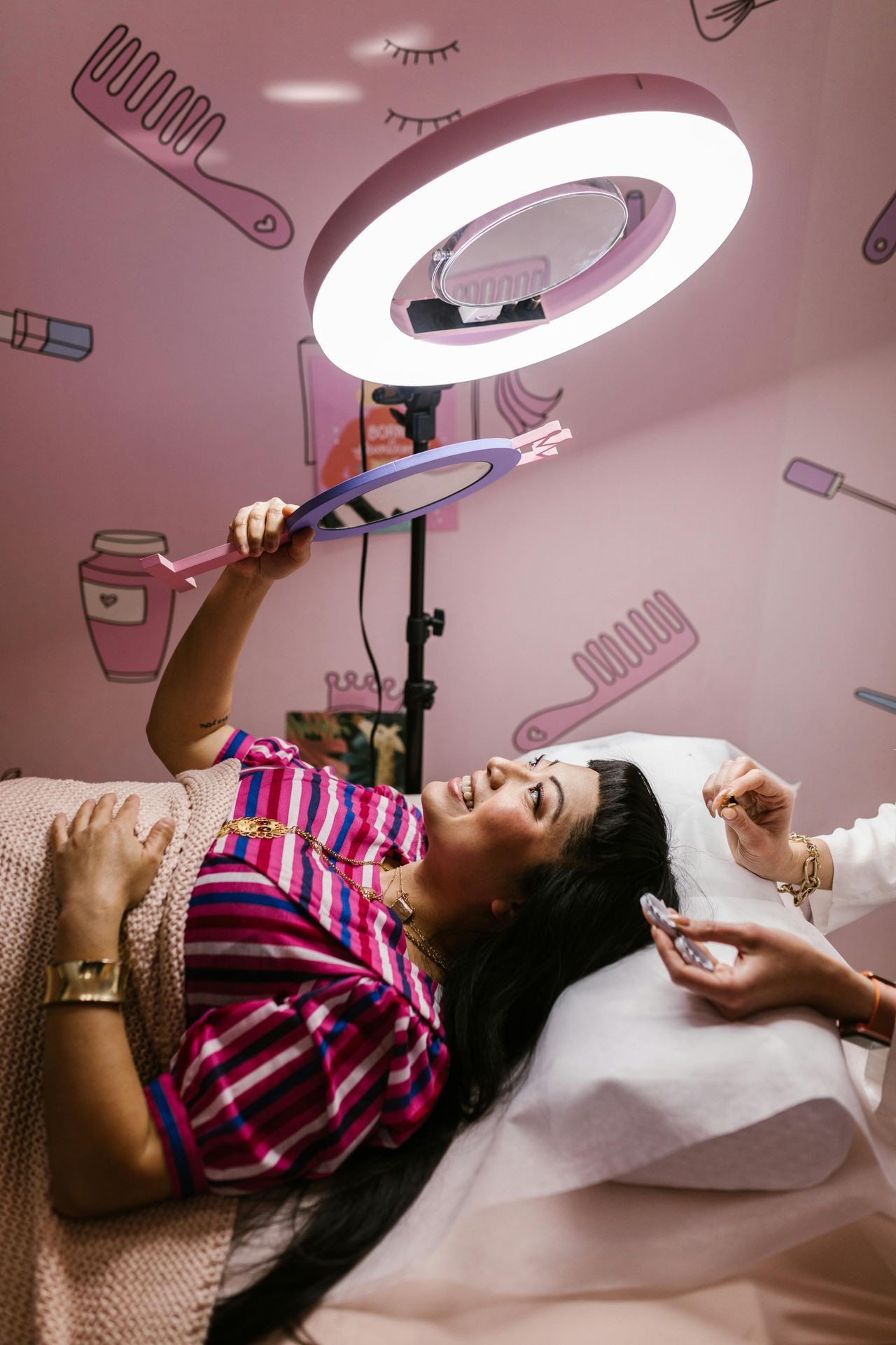 Woman lying under a ring light, holding a mirror, in a beauty salon with pink decor.